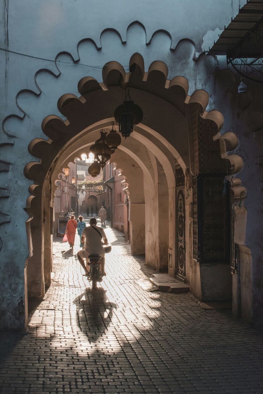A man on a scooter riding through a scalloped archway into a sunlit medina alley